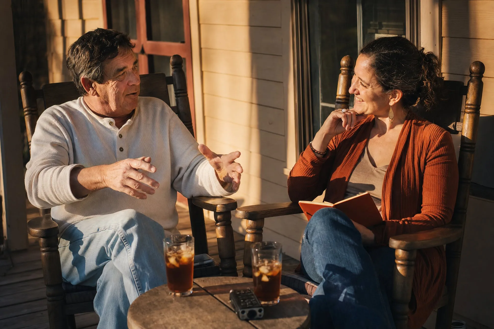 A storyteller and an interviewer in rocking chairs on a porch in golden light, a recorder on the table between them