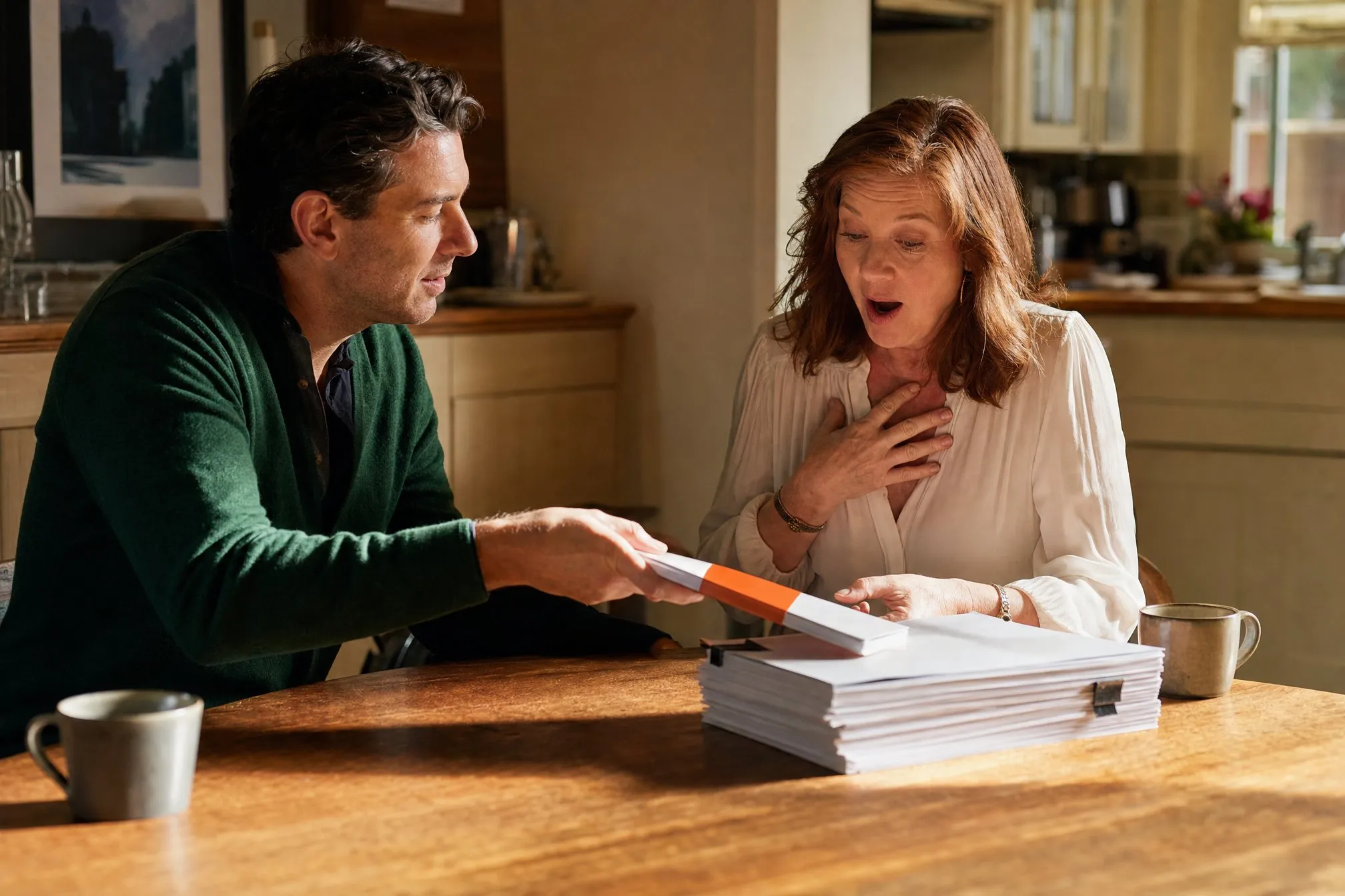 An editor sliding a slim bound proof across a kitchen table to a delighted author, the thick original manuscript beside it